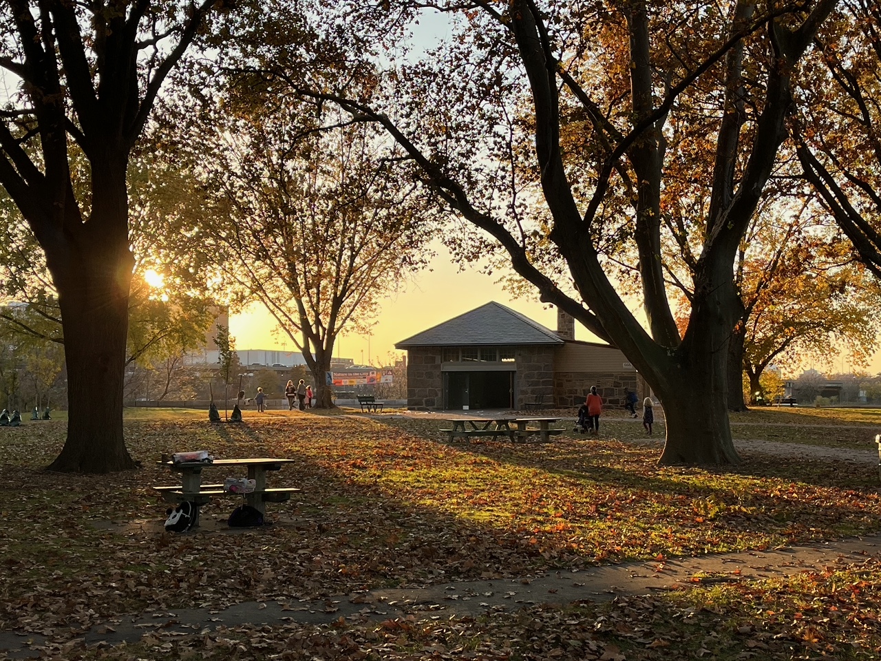 Photo of Magazine Beach and the brick Powder House with the sunset streaming light through the trees and fallen leaves in autumn. People and children are around the picnic tables and the cityscape can be seen across the river.